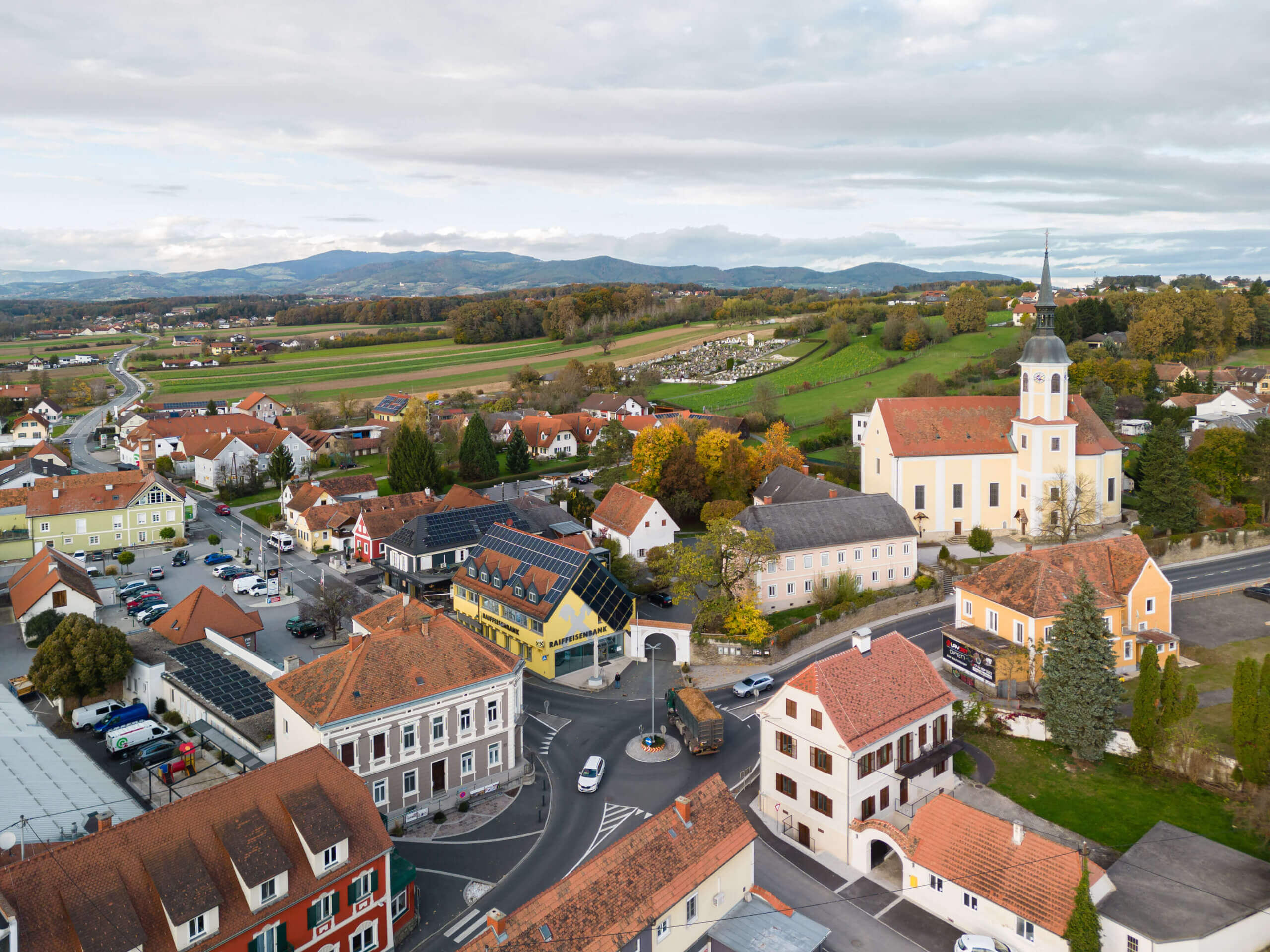 Luftaufnahme eines idyllischen österreichischen Dorfes mit Kirche und Hügeln im Hintergrund, passend für ein regionales Magazin.