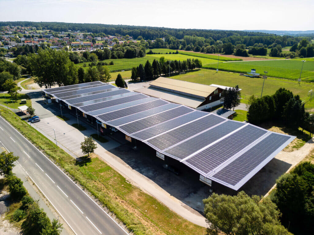 Solardach über Parkplatz und Gebäuden am Stadtrand mit grüner Landschaft im Hintergrund, Perspektive aus der Luft.
