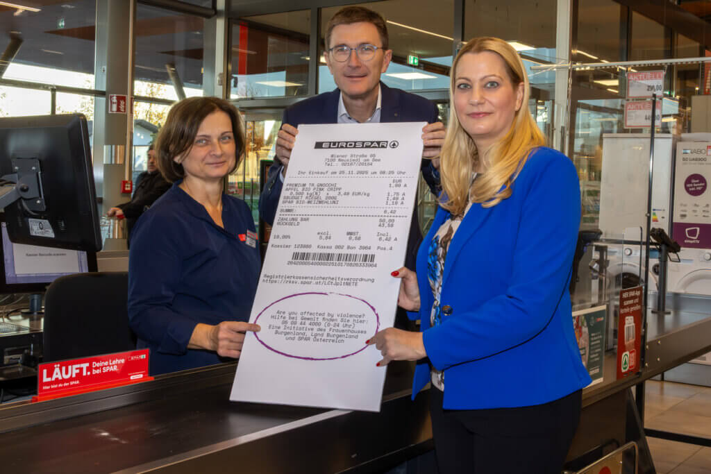 Drei Personen in einem Eurospar zeigen einen übergroßen Kassenbon an einer Kasse des Supermarktes.