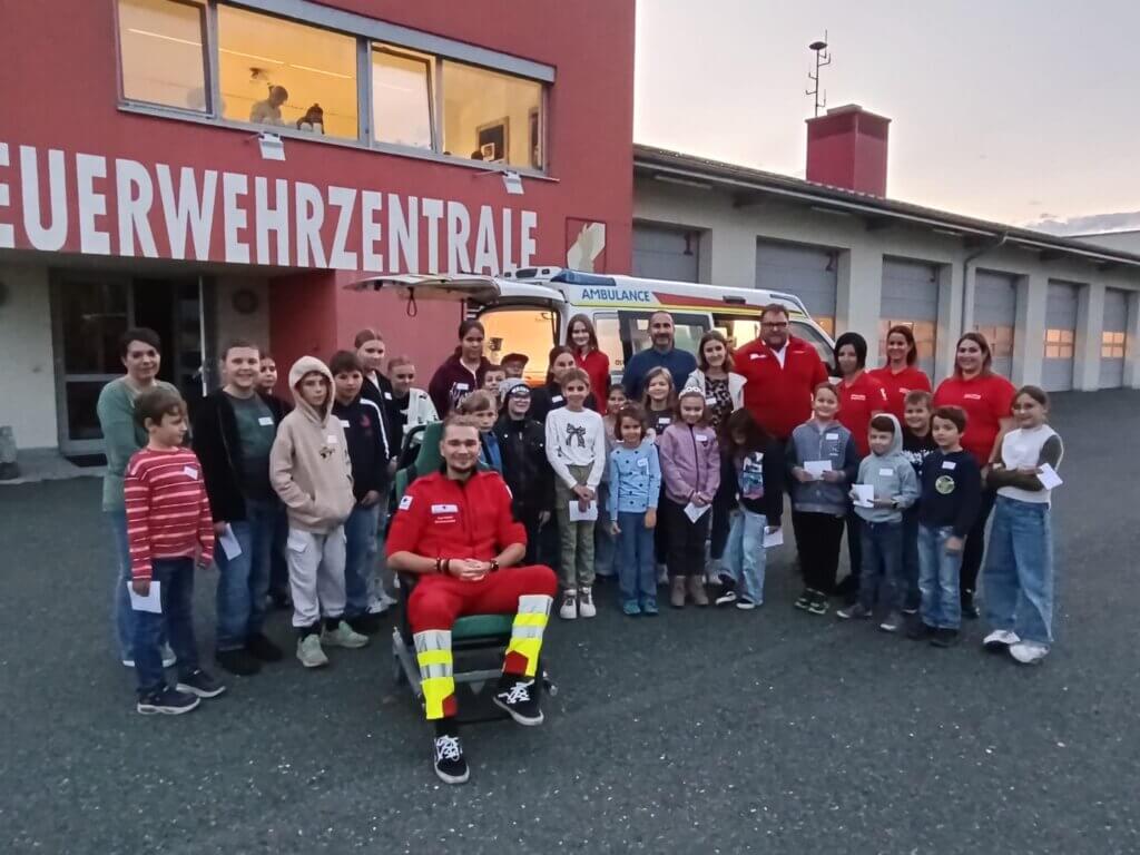 Kinder und Rettungskräfte versammeln sich vor der Feuerwehrenzentrale, Gruppenfoto vor einem Ambulanzfahrzeug bei Abenddämmerung.