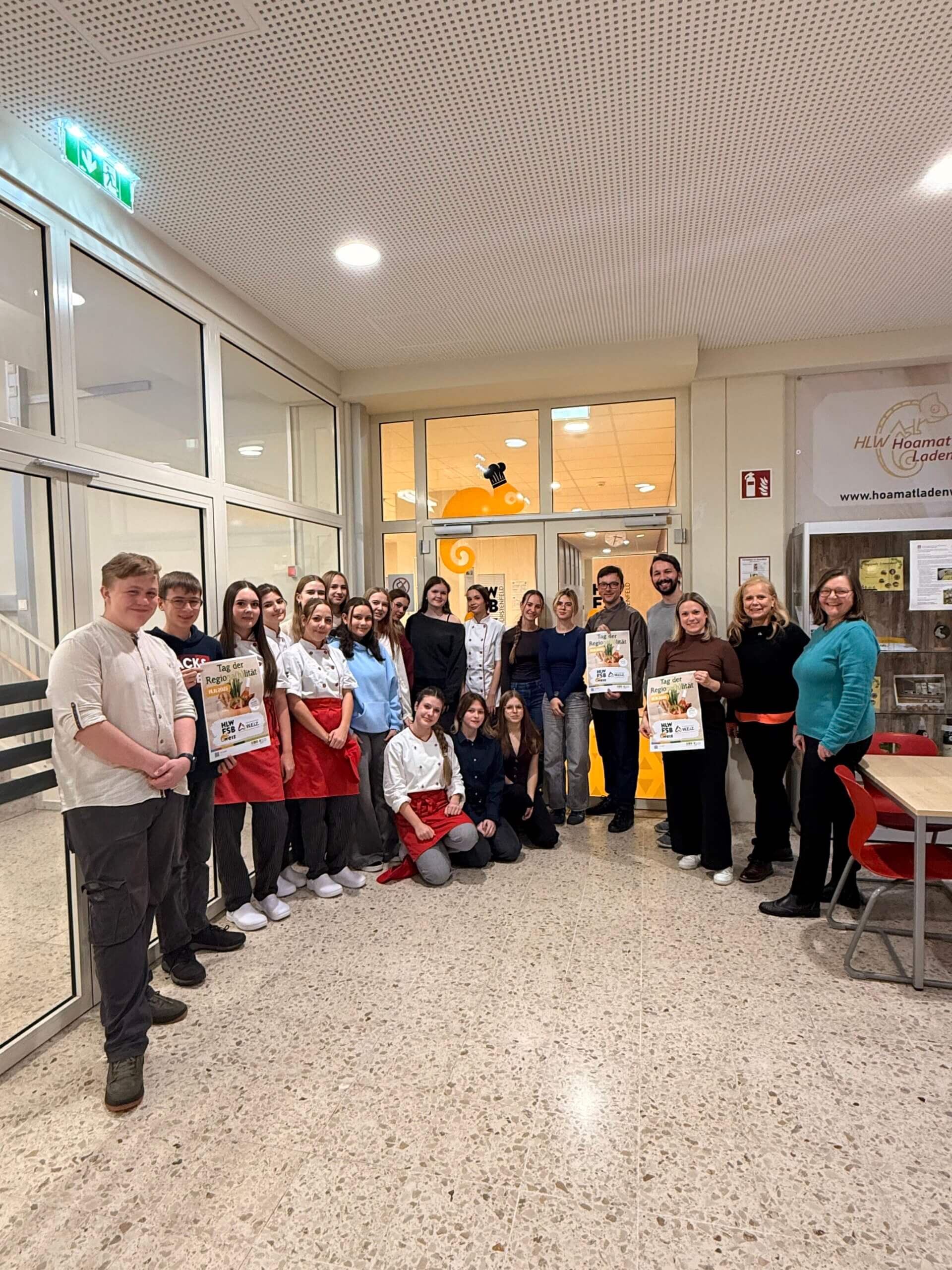 Eine Gruppe von Schülern und Erwachsenen posiert fröhlich gemeinsam mit Plakaten in einem Schulgebäude für ein Foto.