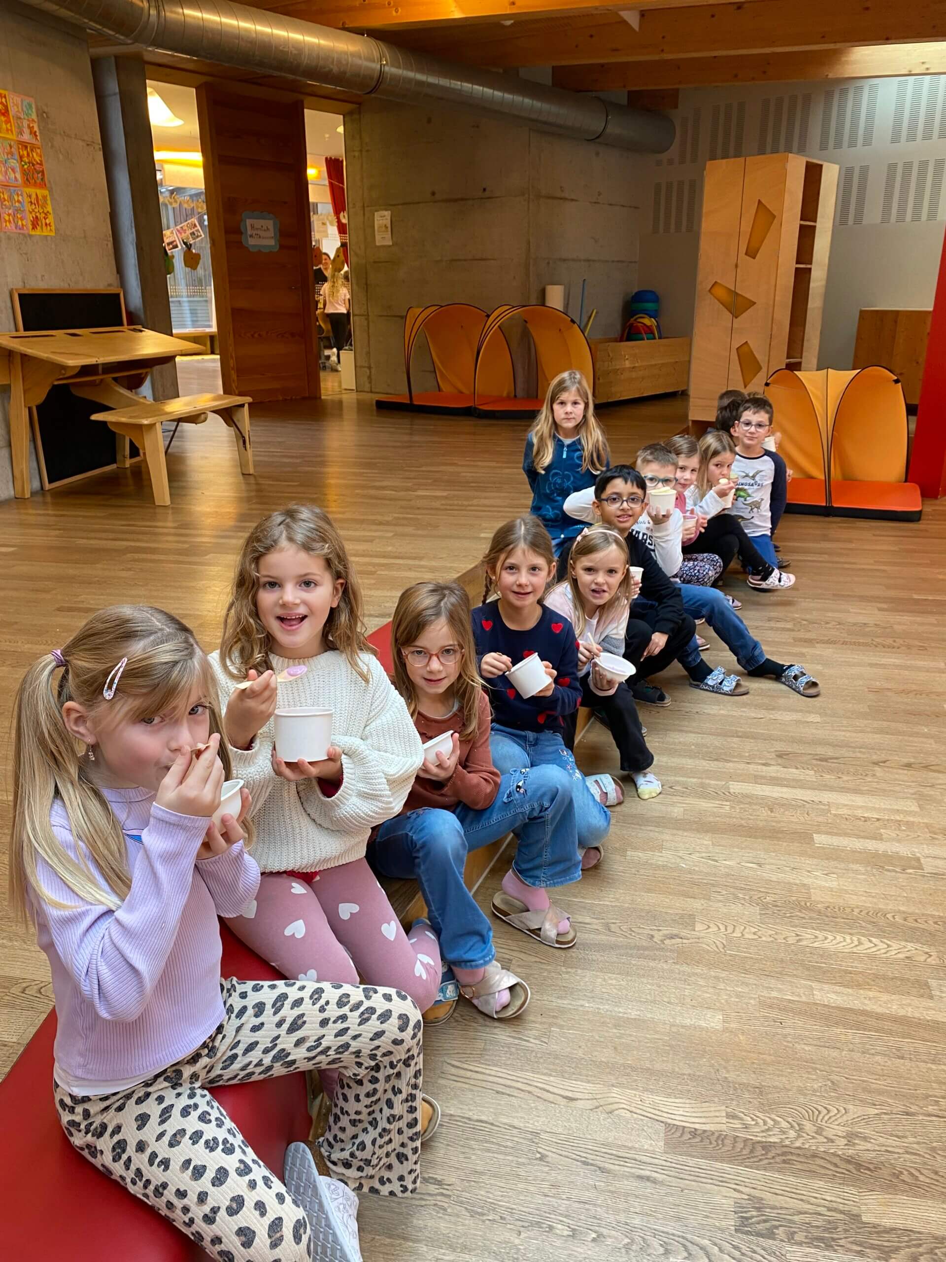 Kinder sitzen in einer Kita, genießen Eisbecher, lächelnd und fröhlich, in einem hellen, einladenden Raum.
