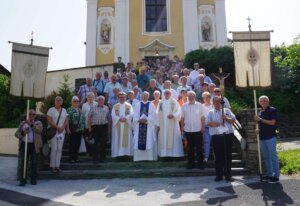 Gruppe von Menschen vor einer Kirche in Feierlaune, begleitet von Priestern in liturgischen Gewändern, aufgenommen bei sonnigem Wetter.