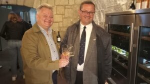 Zwei Männer stoßen mit Weingläsern in einem Weinkeller an, lächelnd, vor einer Steinwand und einem Weinkühler.