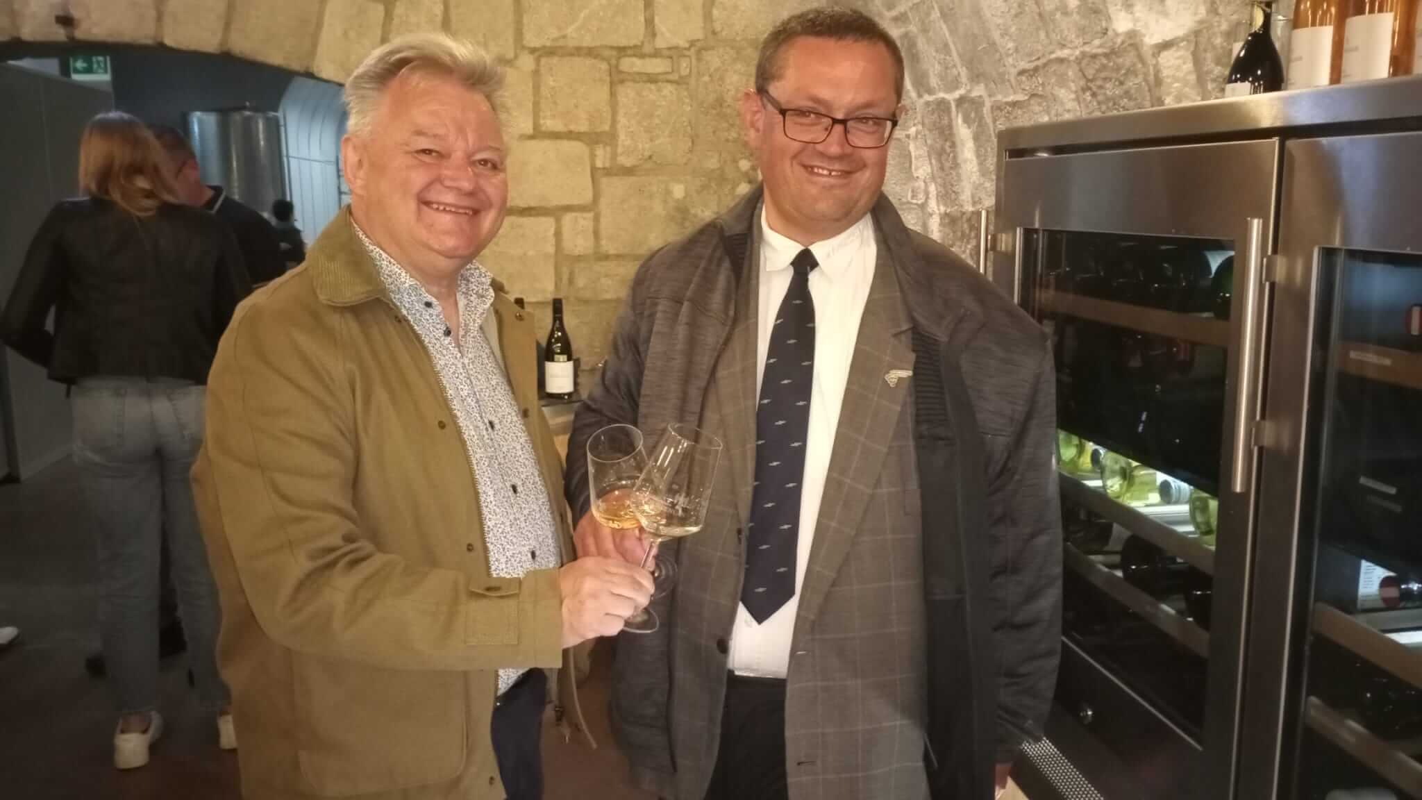 Zwei Männer stoßen mit Weingläsern in einem Weinkeller an, lächelnd, vor einer Steinwand und einem Weinkühler.