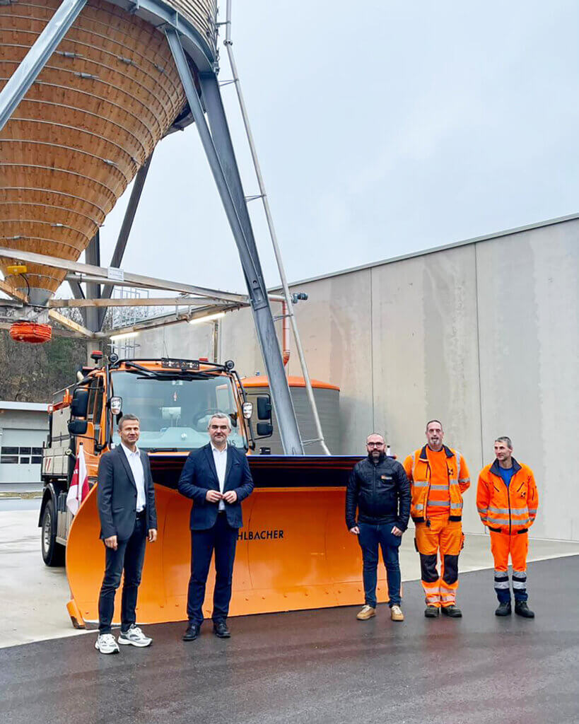 Gruppe von Männern steht vor einem Schneepflug und einem Siloturm, thematisiert regionale Infrastruktur für Prima Magazin.