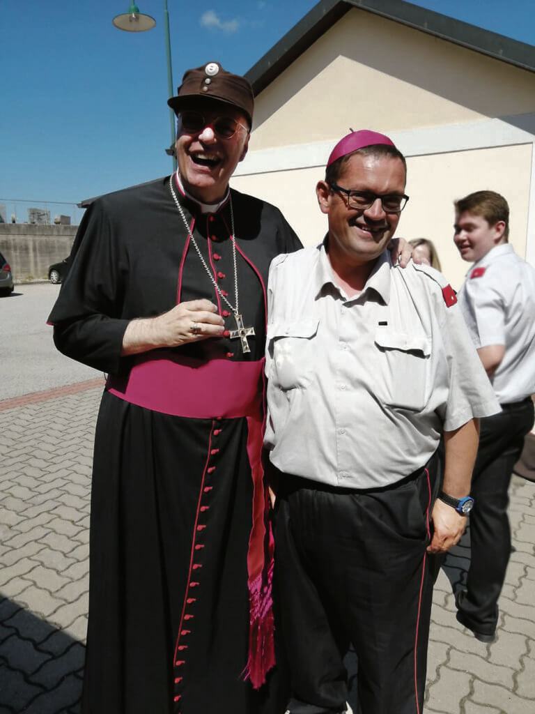 freigegeben_bearb-c-zVg Zwei lächelnde Männer in schwarz-pinker und lässiger Kleidung stehen draußen vor einem Gebäude bei sonnigem Wetter.
