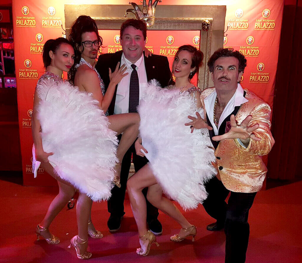 Gruppe von Künstlern und Gästen vor dem Palazzo-Banner, festlich gekleidet, mit Federn und Showkostümen, fröhliche Atmosphäre.