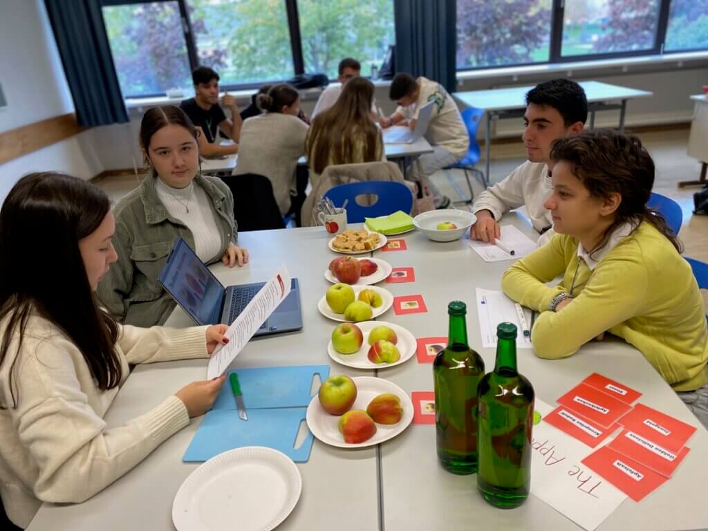 Schüler arbeiten in einer Projektsitzung zu regionalen Apfelsorten, umgeben von Obst und Notizen in einem Klassenraum.