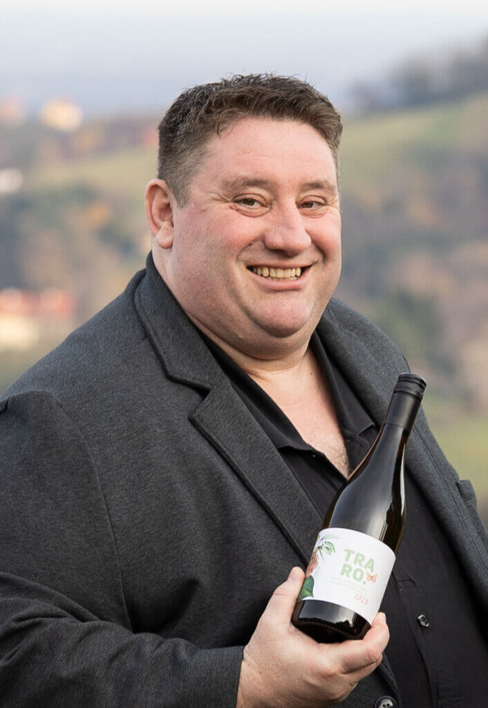 Mann in schwarzem Anzug hält eine Weinflasche vor einer malerischen Landschaft, lächelt freundlich in die Kamera.