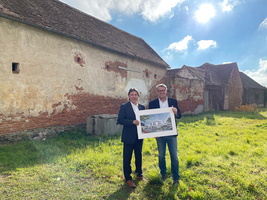 Zwei Männer halten einen Bauplan vor einem alten Gebäude unter blauem Himmel, möglicherweise ein Sanierungsprojekt.