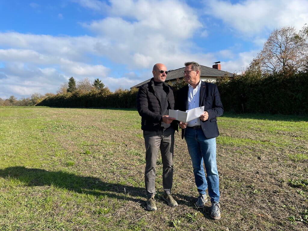 Zwei Männer besprechen Pläne auf einem Feld vor einem Haus unter blauem Himmel, passend für ein regionales Magazin.