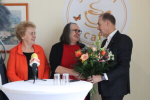 Drei Personen in einem Café, Blumenstrauß und Mikrofon, fröhliche Atmosphäre bei einem regionalen Ereignis für Prima Magazin.