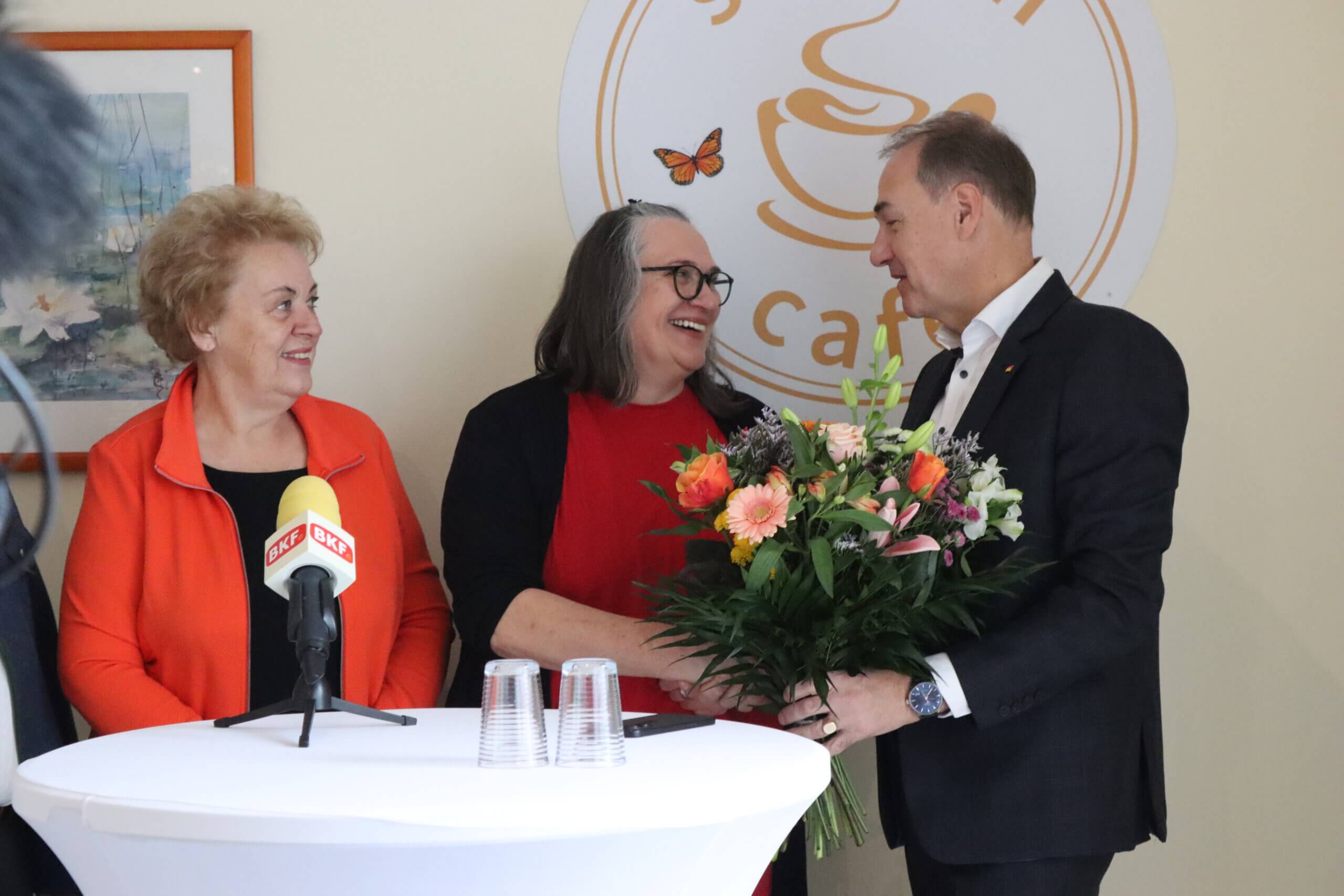 Drei Personen in einem Café, Blumenstrauß und Mikrofon, fröhliche Atmosphäre bei einem regionalen Ereignis für Prima Magazin.
