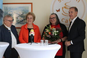 Eine Gruppe von vier Personen bei einer Veranstaltung, eine Frau hält einen Blumenstrauß. Mikrofon mit „BKF“-Logo sichtbar.