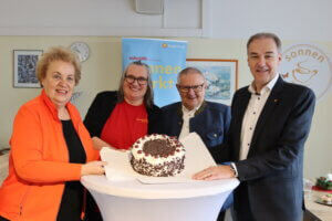 Vier lächelnde Personen präsentieren eine Schwarzwälder Kirschtorte bei einer Veranstaltung mit einem Banner im Hintergrund.