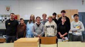 Eine Gruppe Jugendlicher und ein Lehrer in einem Klassenzimmer, lächelnd für ein regionales Magazin-Foto.