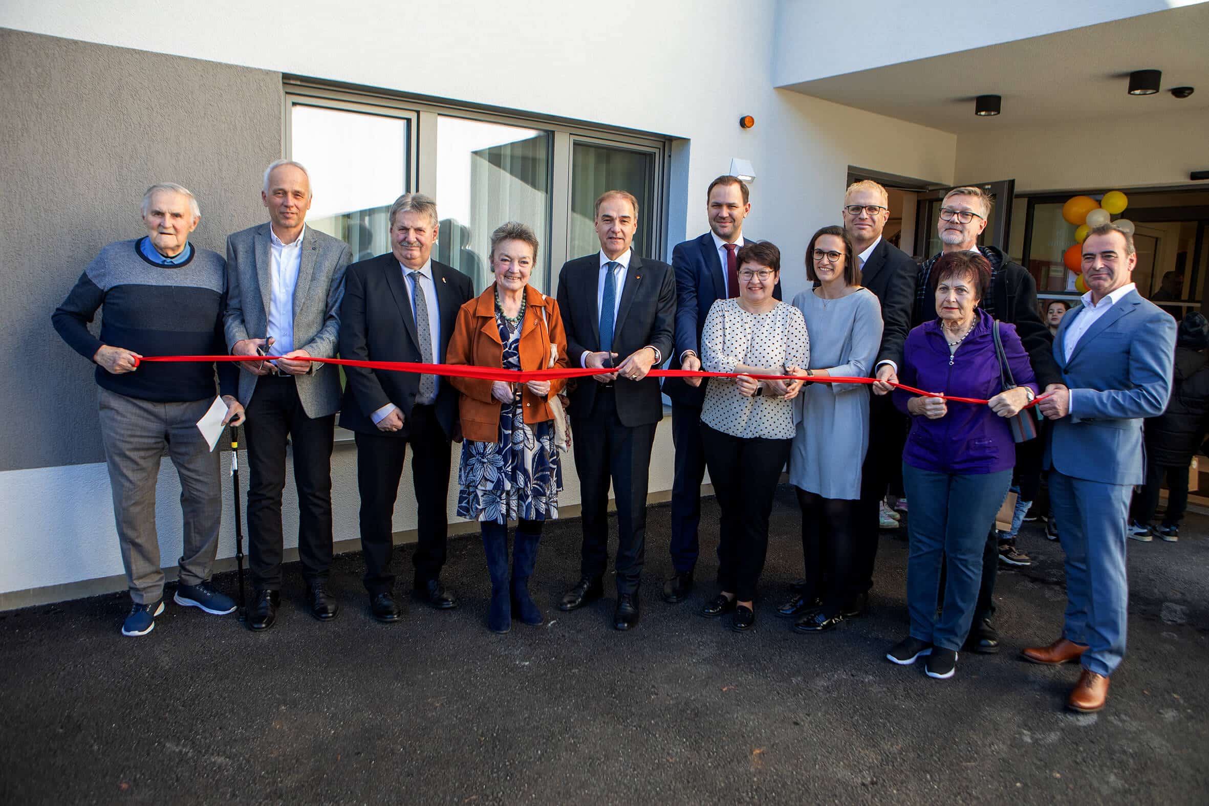 Gruppe von Menschen durchschneidet feierlich ein rotes Band vor einem neuen Gebäude, als Teil einer Eröffnung.
