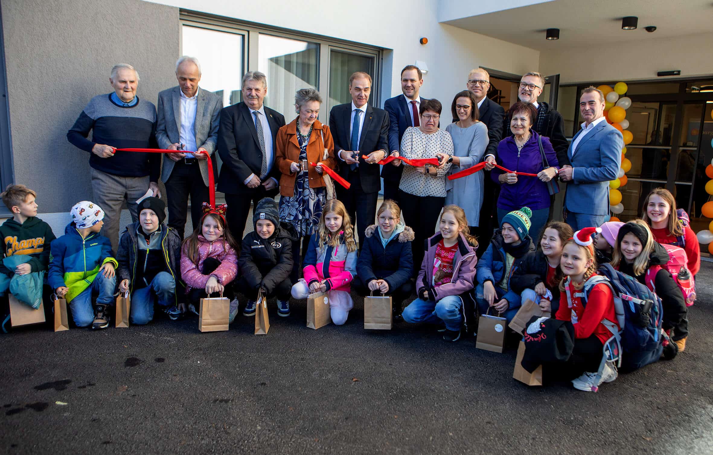 Gruppeneröffnung mit Erwachsenen und Kindern vor einem Gebäude. Feierliche Eröffnung mit rotem Band und fröhlicher Stimmung.