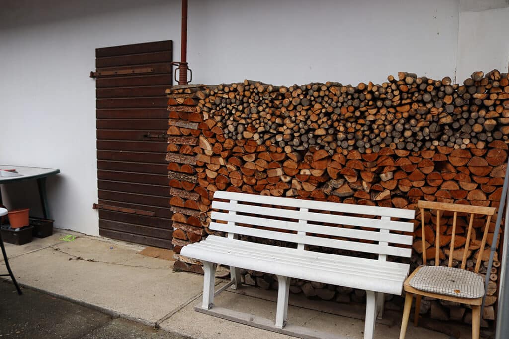 Holzstapel an weißer Wand mit weißer Bank und Holzstuhl; einladende, rustikale Gartenszene für regionales Magazin.