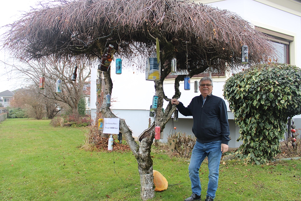 Ein Mann steht neben einem Baum mit aufgehängten Bierflaschen vor einem Haus, im Rahmen eines humorvollen Winterarrangements.
