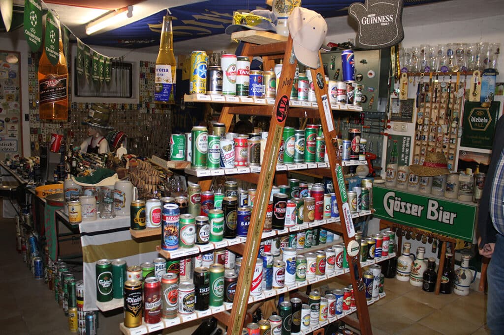 Bunte Bierdosen-Sammlung auf hölzernem Regal in privater Bar mit Bierkrügen und Deko, ideal für Brauerei-Liebhaber.