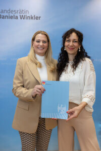 Zwei Frauen lächeln vor einem blauen Hintergrund mit einem Dokument des Landes Burgenland in der Hand für Prima Magazin.