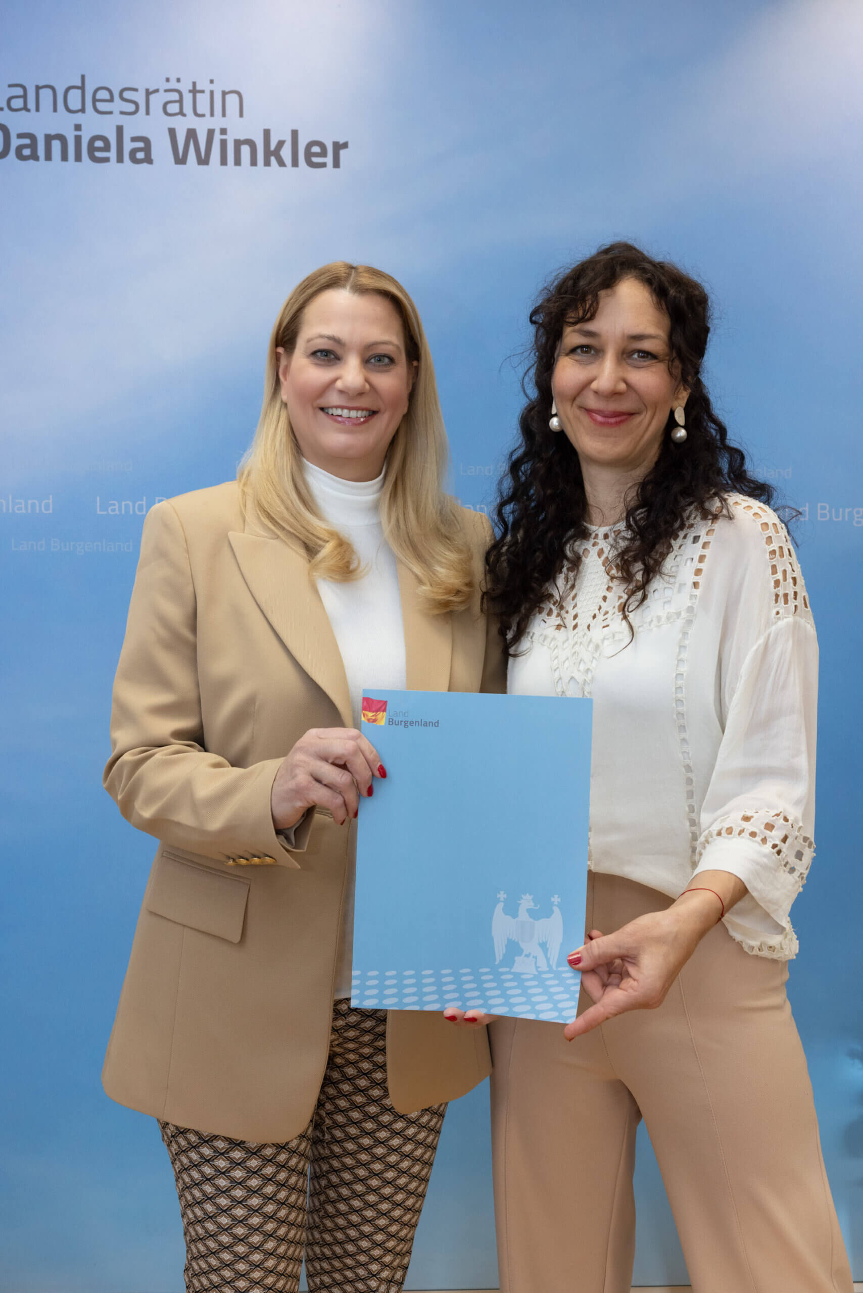 Zwei Frauen lächeln vor einem blauen Hintergrund mit einem Dokument des Landes Burgenland in der Hand für Prima Magazin.