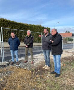 Vier Männer stehen auf einer Baustelle, betrachten das Gelände hinter einem Zaun; blauer Himmel im Hintergrund.