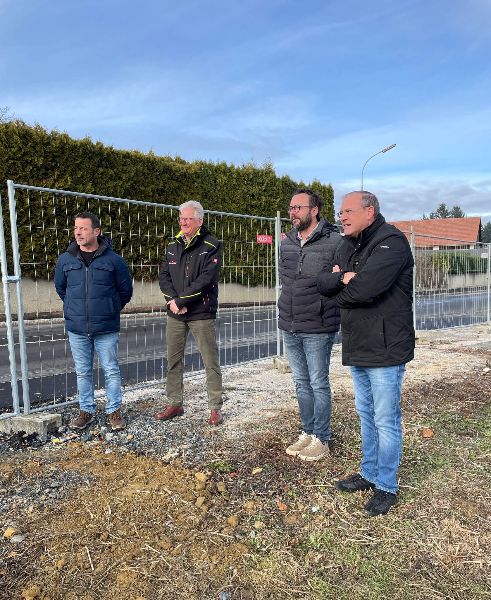 Vier Männer stehen auf einer Baustelle, betrachten das Gelände hinter einem Zaun; blauer Himmel im Hintergrund.