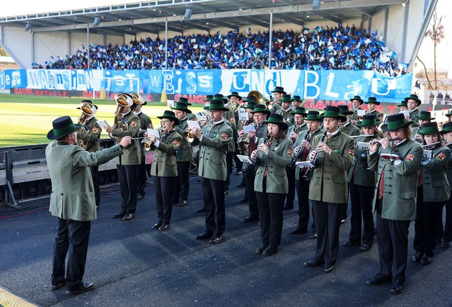 Blaskapelle in traditioneller Tracht spielt bei einem Fußballspiel, eine voll besetzte Tribüne mit Fans im Hintergrund.