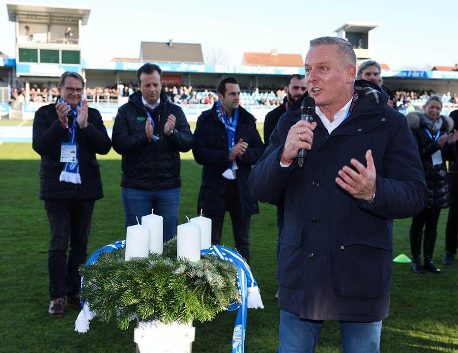 Mann hält Rede bei Outdoor-Event neben Adventskranz, umgeben von applaudierenden Personen auf Sportplatz des Prima Magazins.