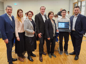 Personengruppe in formeller Kleidung in einem Büro steht zusammen, eine Person hält einen Laptop mit einem geöffneten Programm.