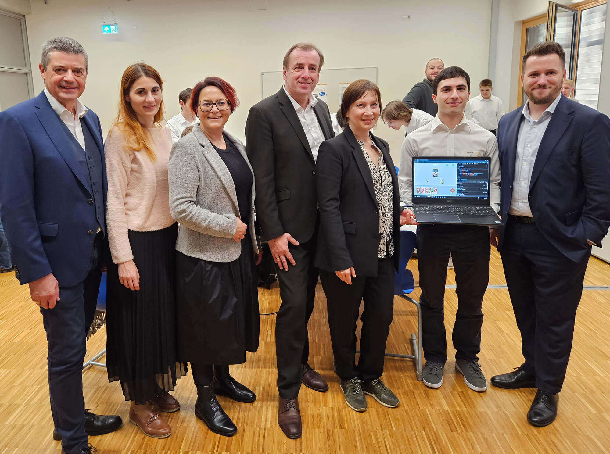 Personengruppe in formeller Kleidung in einem Büro steht zusammen, eine Person hält einen Laptop mit einem geöffneten Programm.
