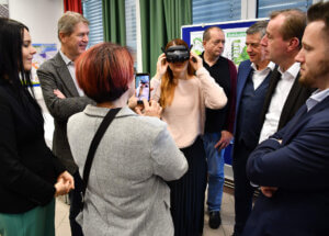 Eine Frau testet VR-Brille, umgeben von interessiertem Publikum, während eine andere Person Fotos aufnimmt.