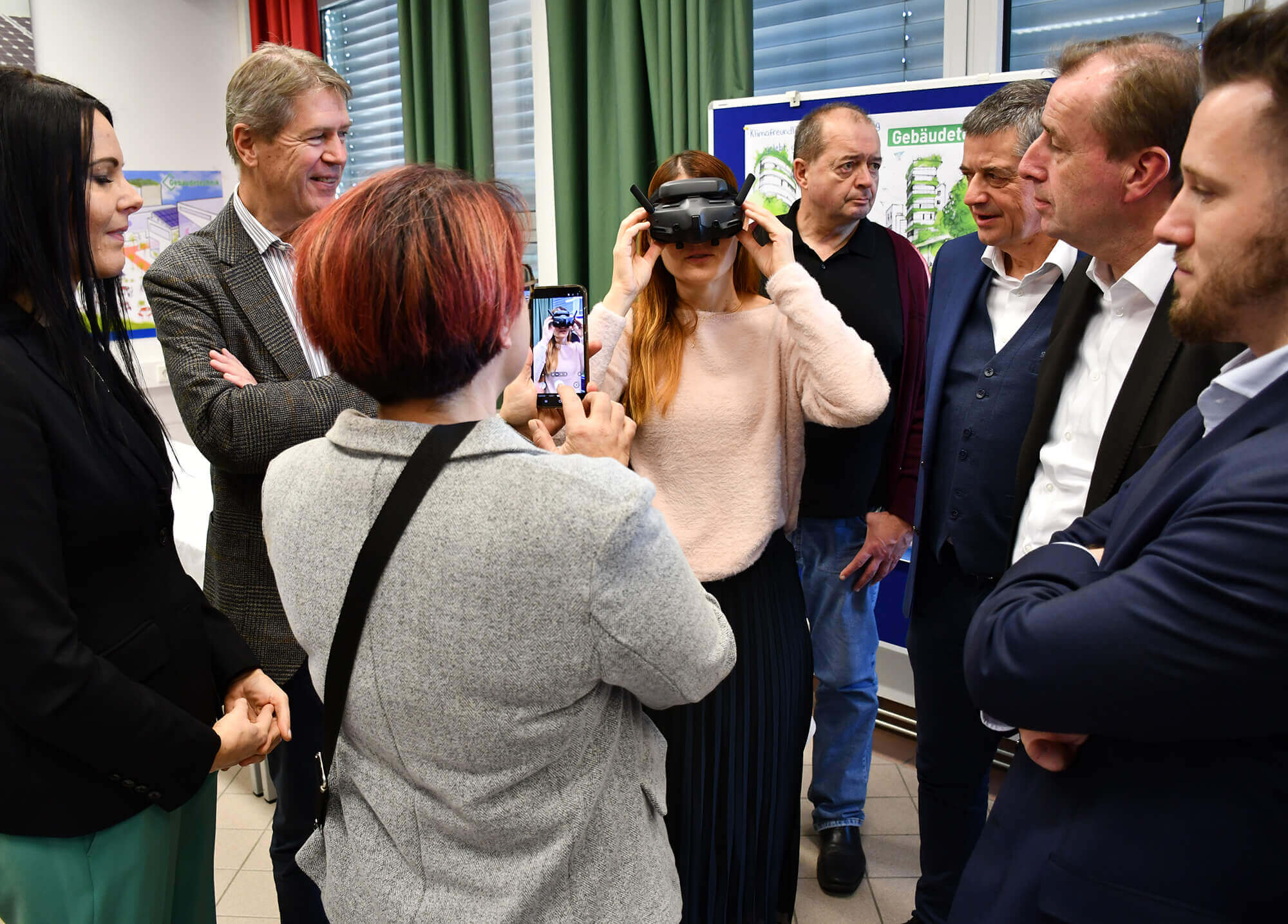 Eine Frau testet VR-Brille, umgeben von interessiertem Publikum, während eine andere Person Fotos aufnimmt.