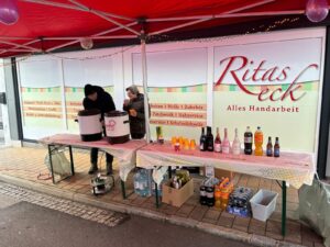 Zwei Personen servieren Getränke unter einem roten Zelt bei Ritas Eck, einem Geschäft für Handarbeiten, auf einem Straßenfest.