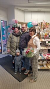 Familie im Geschäft mit Blumenstrauß, umgeben von Produkten und Luftballons, feiert ein lokales Ereignis für Prima Magazin.