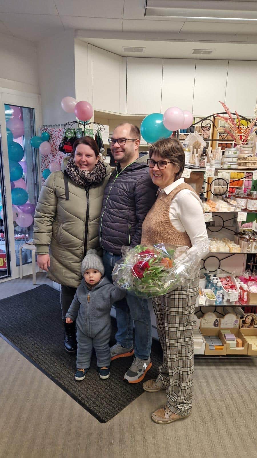 Familie im Geschäft mit Blumenstrauß, umgeben von Produkten und Luftballons, feiert ein lokales Ereignis für Prima Magazin.