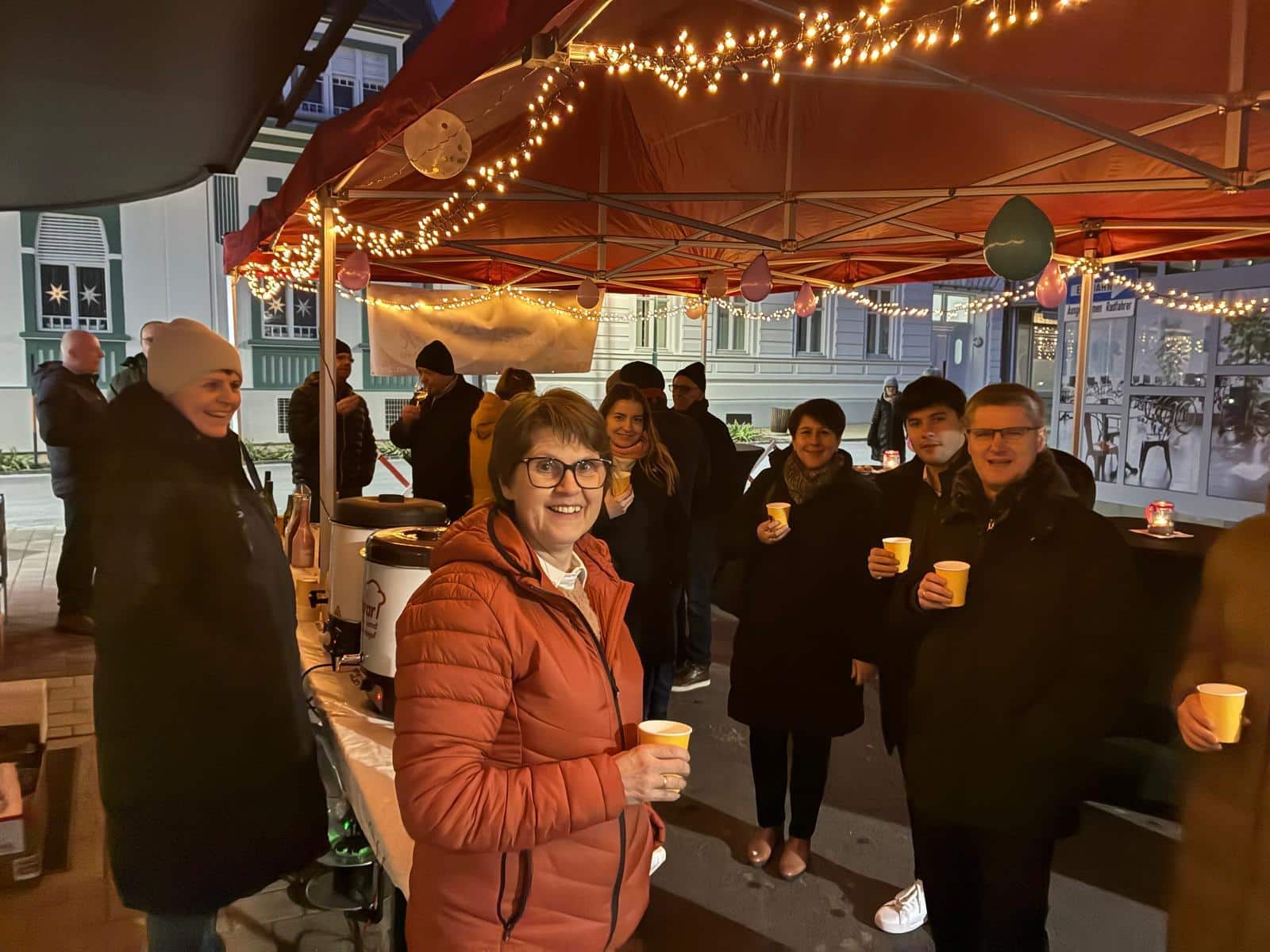 Eine fröhliche Gruppe genießt heiße Getränke unter einem festlich beleuchteten Stand auf einem regionalen Wintermarkt.
