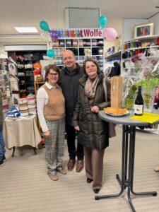 Personengruppe bei einem Wollegeschäft-Event, umgeben von Garn und Strickartikeln, mit Snacks und Getränken auf einem Tisch.