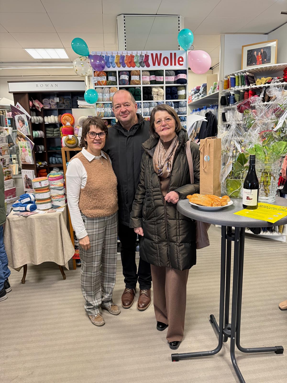 Personengruppe bei einem Wollegeschäft-Event, umgeben von Garn und Strickartikeln, mit Snacks und Getränken auf einem Tisch.
