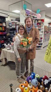 Personen in einem Wollgeschäft mit Blumenstrauß und Garn im Vordergrund, gemeinsames Lächeln zeigt lokale Gemeinschaftlichkeit.