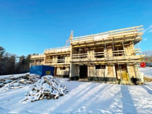 Schneebedeckte Baustelle eines zweistöckigen Gebäudes mit Gerüst, blauer Container und Baumaterialien unter klarem Himmel.