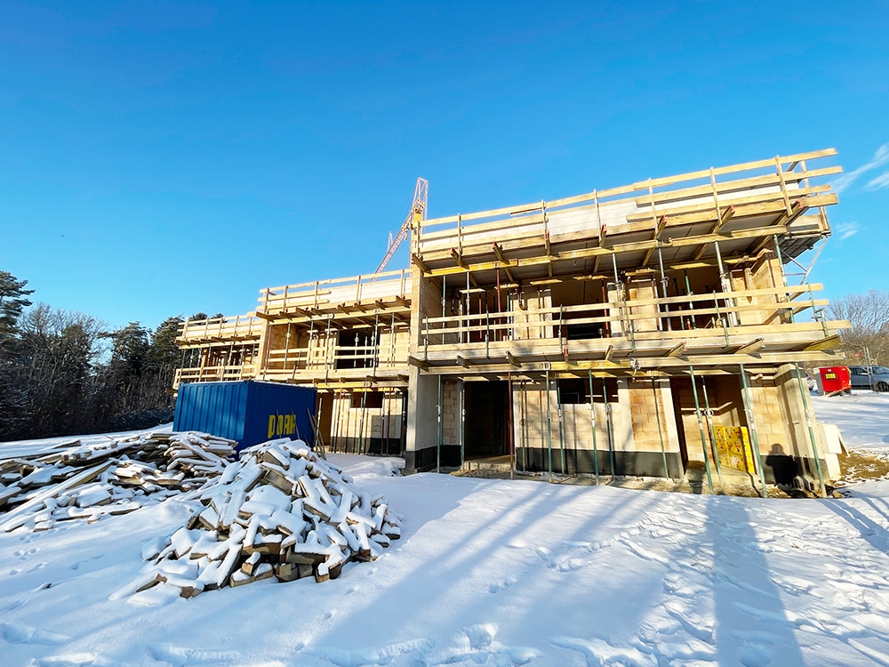 Schneebedeckte Baustelle eines zweistöckigen Gebäudes mit Gerüst, blauer Container und Baumaterialien unter klarem Himmel.