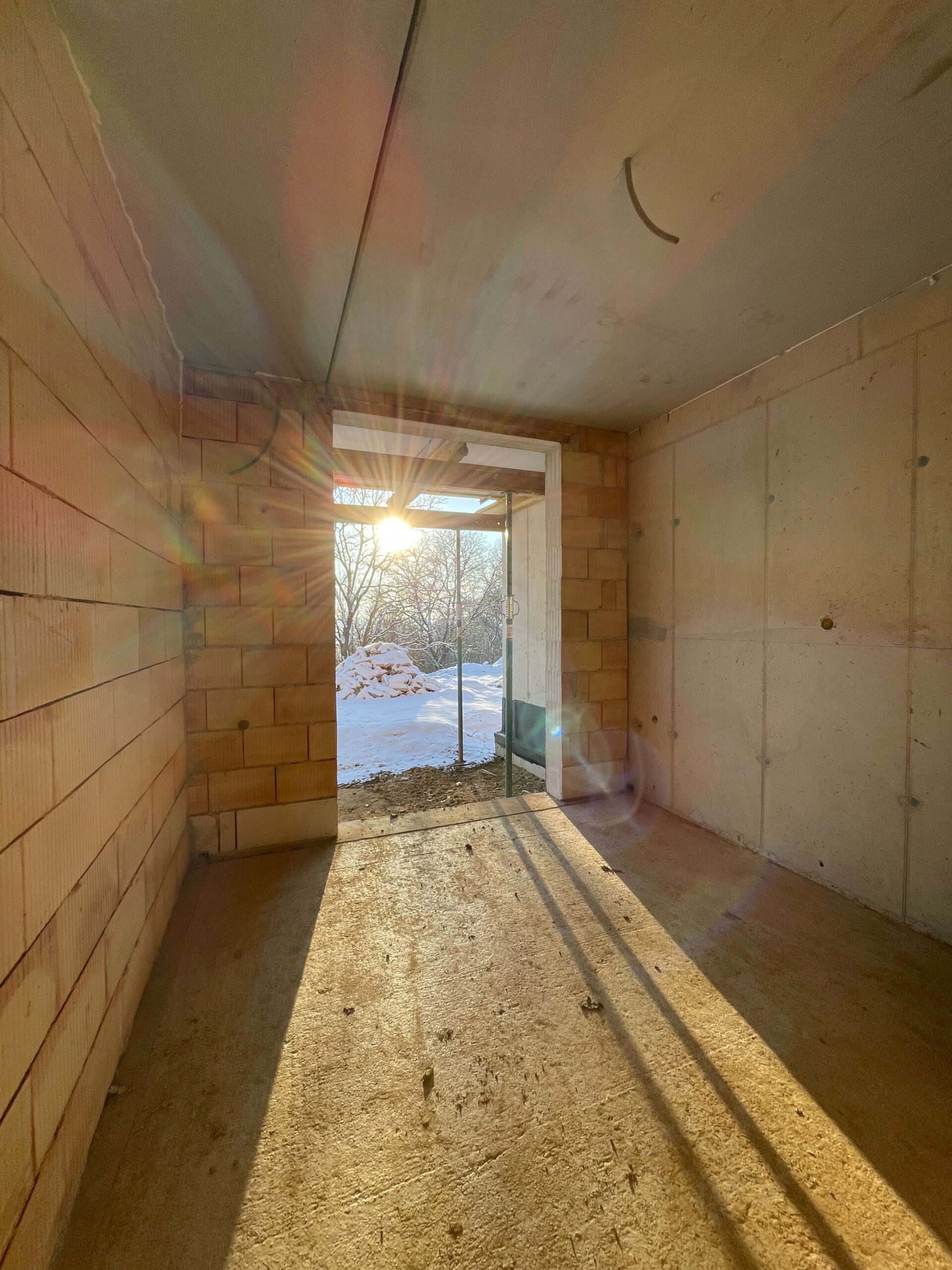 Blick durch einen Rohbau bei Sonnenaufgang, das ein winterliches Landschaftsbild mit Schneedecke im Hintergrund enthüllt.