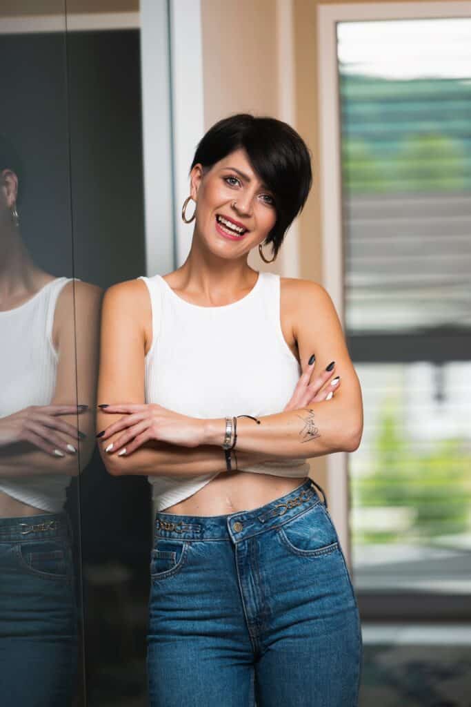 Junge Frau mit kurzem Haar und tätowierten Armen lächelt entspannt in die Kamera, vor einem Fenster, für Prima Magazin.