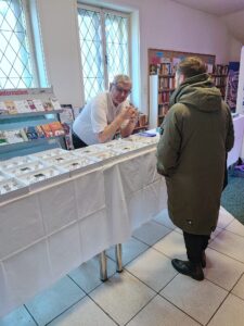 Mann berät einen Besucher an einem Schmuckstand auf einer Messe; Bücherregale im Hintergrund, informative Atmosphäre im Prima Magazin.