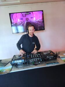 DJ spielt Musik auf einer Veranstaltung mit Burg Güssing im Hintergrund, präsentiert von Prima Magazin auf einem Bildschirm.