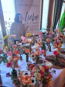 Verzierte Tafel voller bunter Blumen, begleitet von einer Frau, vor einem Banner mit dem Schriftzug Liebe auf den ersten Blick.